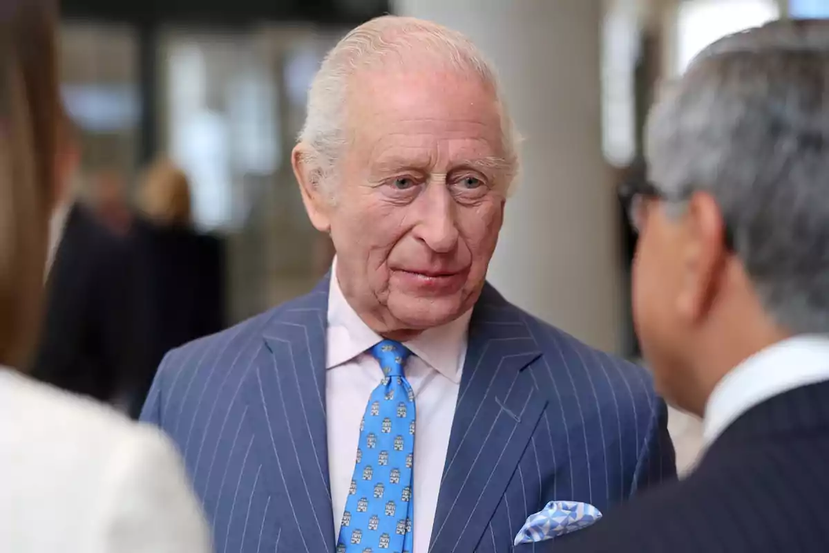 Charles III in a blue striped suit talks with another person in an indoor setting. Charles III in a blue striped suit talks with another person in an indoor setting.