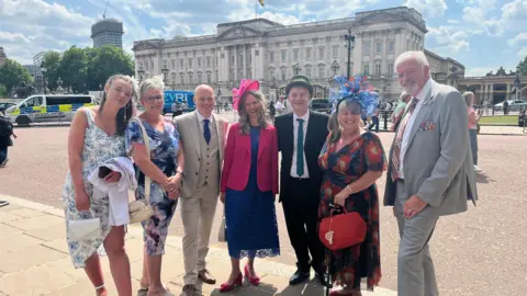 Karl Bird/ BBC Left to Right: Emma Sellars, Ms Sellars' mother, Stuart Wills, Mr Wills' partner Karen, Scott Dalton, Liz Quiney and Ms Quiney's partner. They are all dressed in smart clothes and are stood in front of Buckingham Palace.