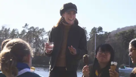 Kensington Palace Princess Kate stands smiling and holding a glass of water in front of a late. Girls are sat at a table in front of her eating ice creams and smiling. 