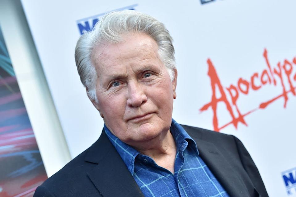 Person wearing a blue plaid shirt and dark blazer at an event with "Apocalypse" text on backdrop