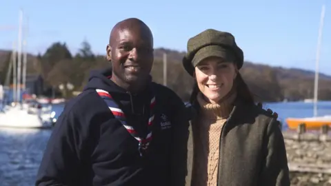 Kensington Palace Princess Kate stands smiling with his arm around a man. He is wearing a scouts tie and a navy jumper. They are standing in front of a lake. 