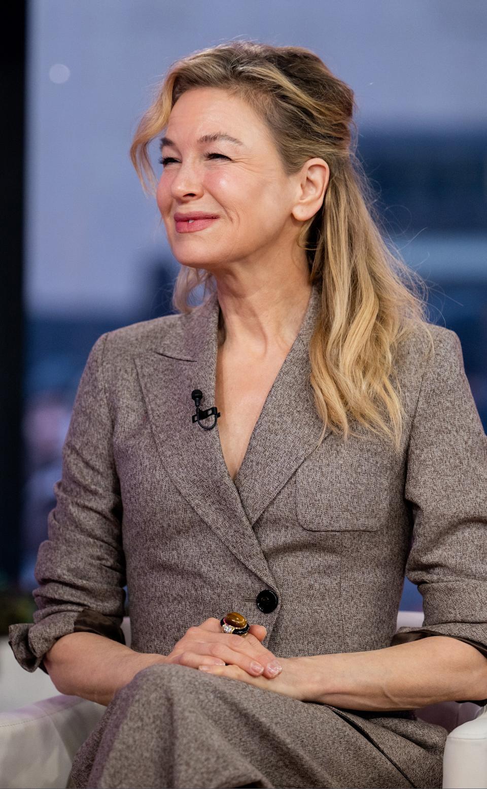 Renée Zellweger smiling in a tailored suit during an interview