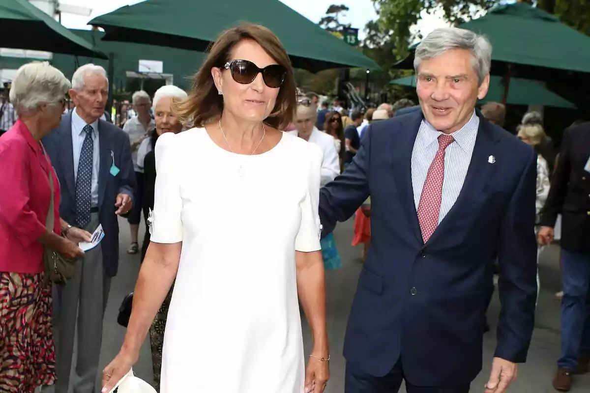 A well-dressed couple walks outdoors surrounded by other people at a social event. A well-dressed couple walks outdoors surrounded by other people at a social event.