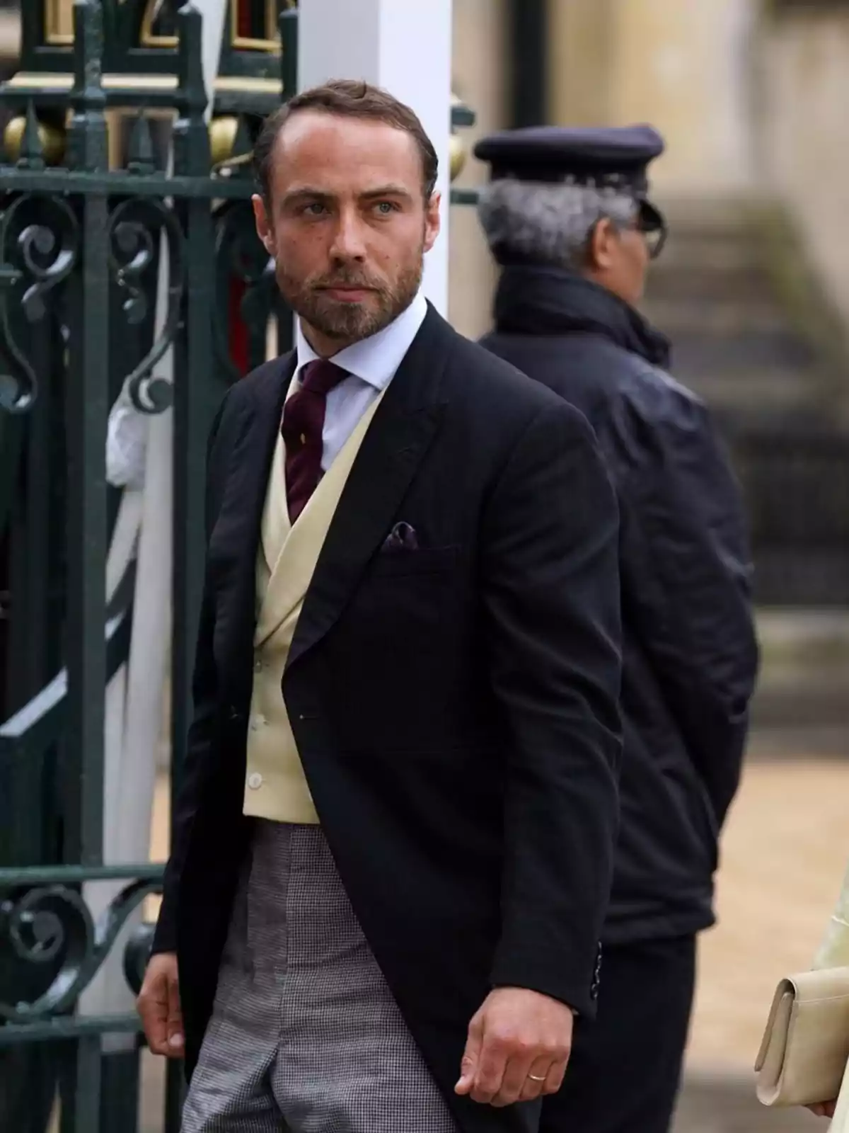 A bearded man with short hair, dressed in a formal suit with a black jacket, beige vest, red tie and checked trousers, is standing in front of a green fence, while a person in a dark uniform and cap stands with his back turned in the background.