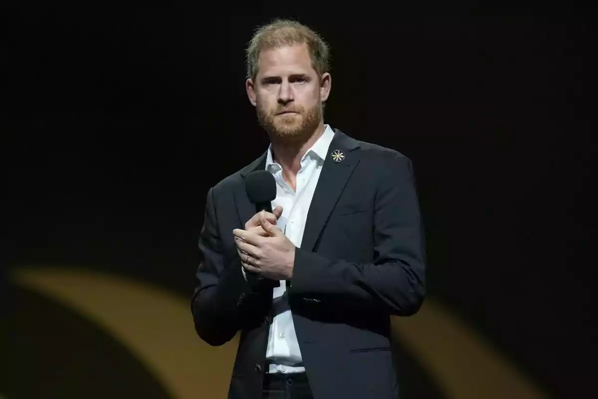 A man with a beard and short hair holds a microphone while wearing a dark suit and a white jersey. A man with a beard and short hair holds a microphone while wearing a dark suit and a white jersey.