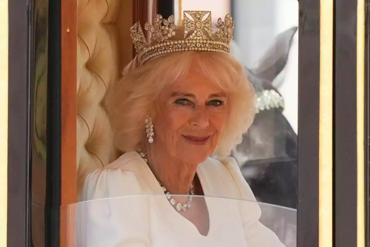 A woman with blonde hair and a jewel-adorned crown, dressed in white, smiles from inside a carriage.
