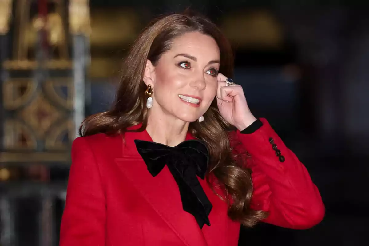 Kate Middleton, wearing a red coat with a black bow, smiles while touching her ear. Kate Middleton, wearing a red coat with a black bow, smiles while touching her ear.