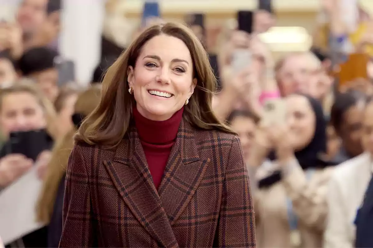 Kate Middleton smiling in a brown plaid coat and a dark red sweater, surrounded by people holding mobile phones.