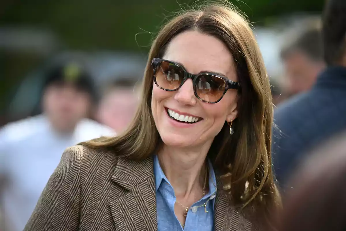 Kate Middleton smiling with sunglasses and long hair, wearing a brown blazer and a blue jersey, in an outdoor setting.