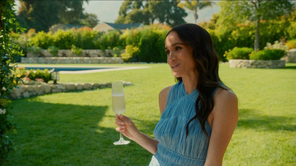 Meghan Markle holding a champagne flute in a backyard setting.