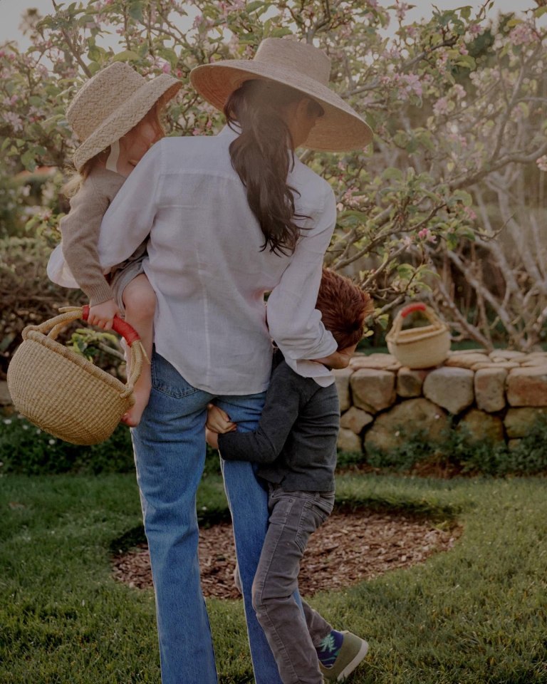 Woman carrying a young child while another child holds onto her legs in a garden.