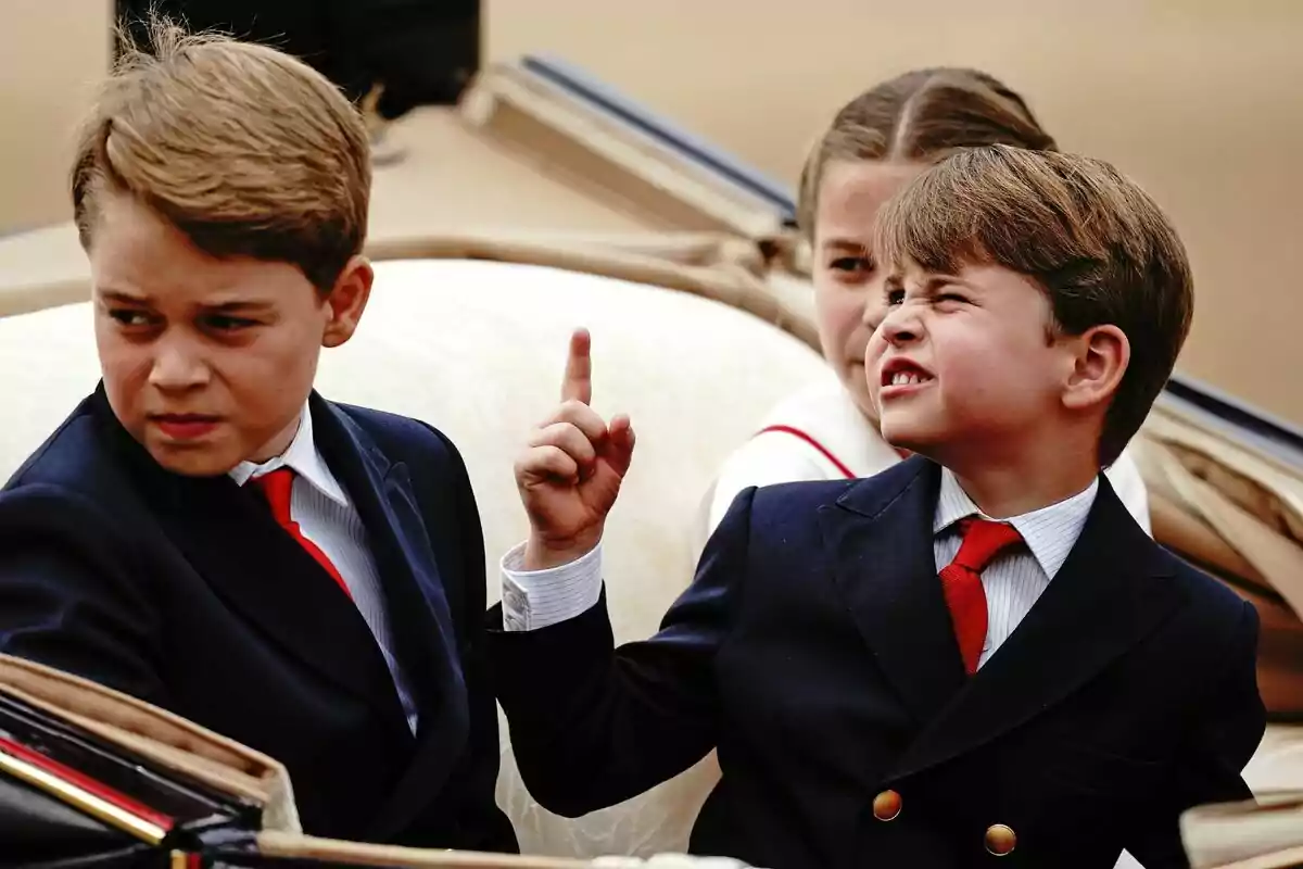 The formally dressed Princes George, Louis and Charlotte are sitting in a carriage, Louis is raising his index finger while grimacing.