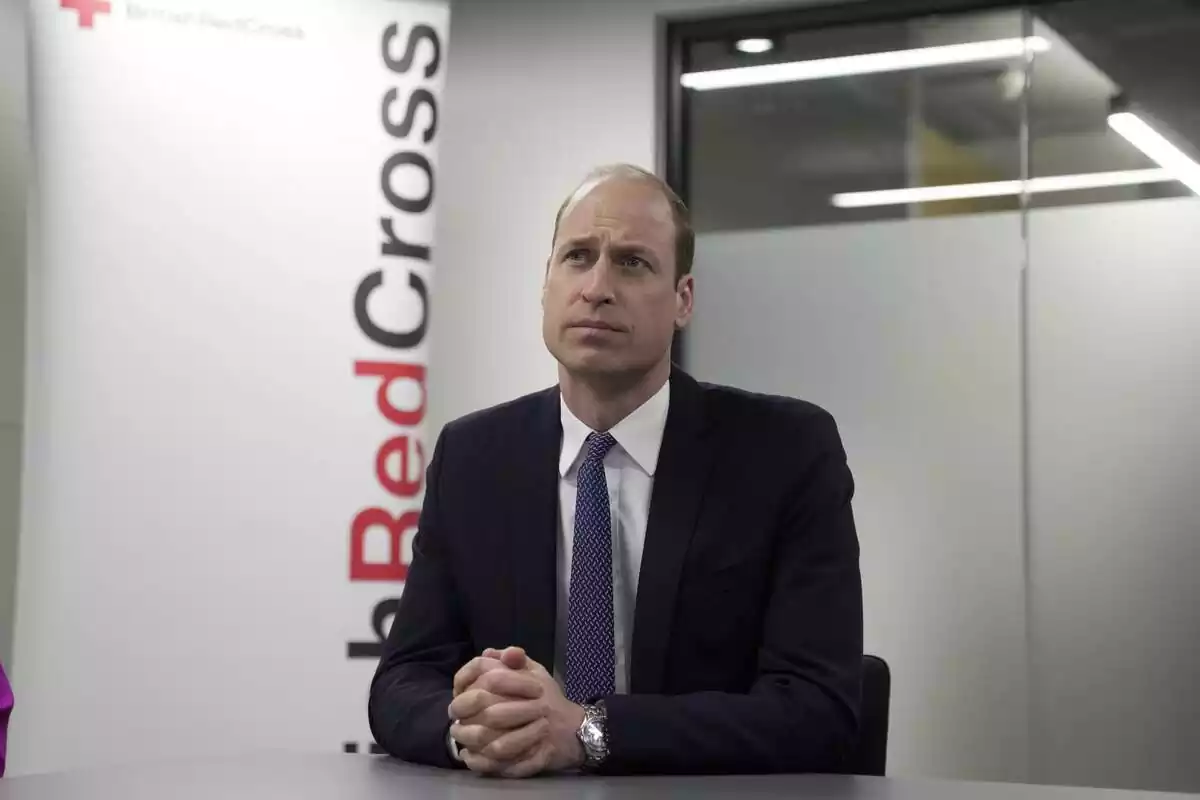 Prince William serious in black suit and blue tie, hands together and sitting