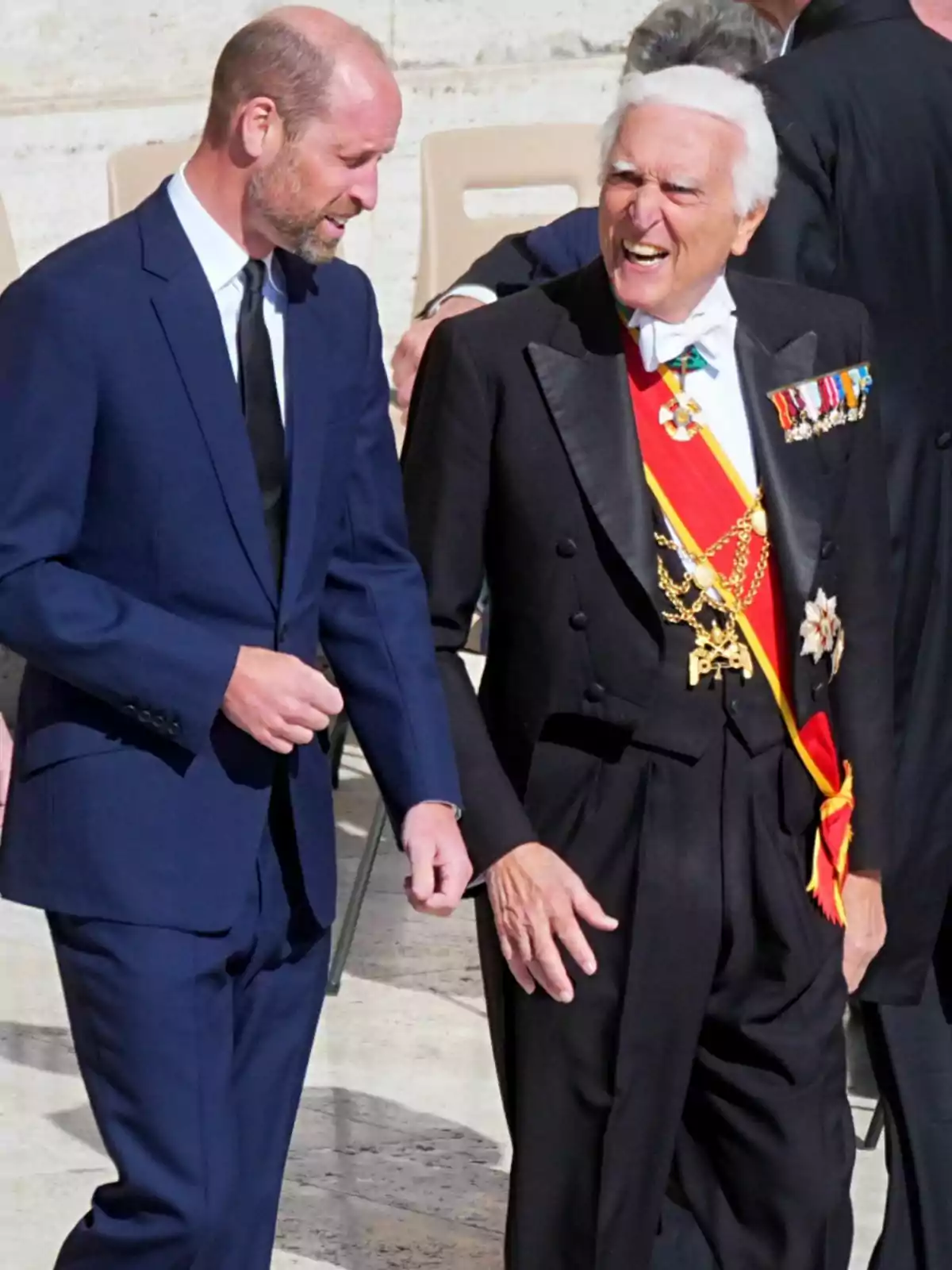 Prince William and another man are having an animated conversation; one is wearing a blue suit and the other a formal suit with decorations and a red sash. Prince William and another man are having an animated conversation; one is wearing a blue suit and the other a formal suit with decorations and a red sash.
