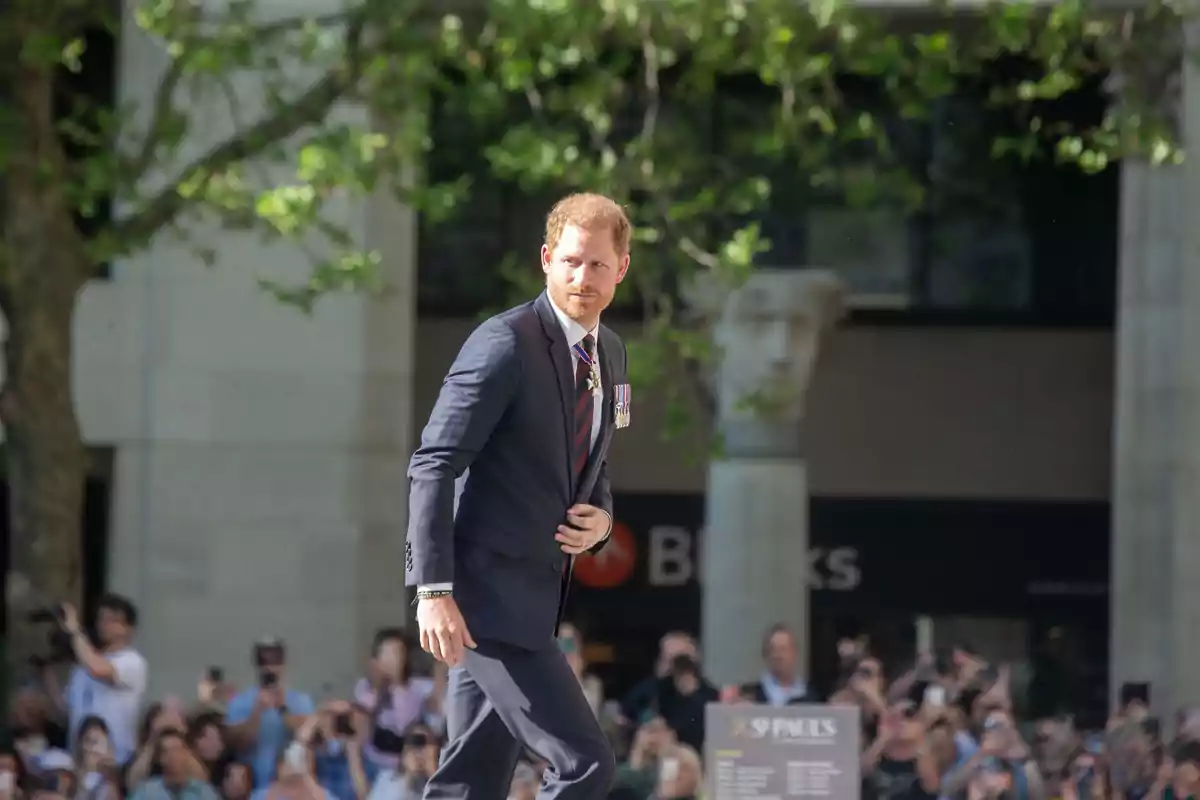 Prince Harry in a dark suit walks outdoors while being photographed by a crowd.