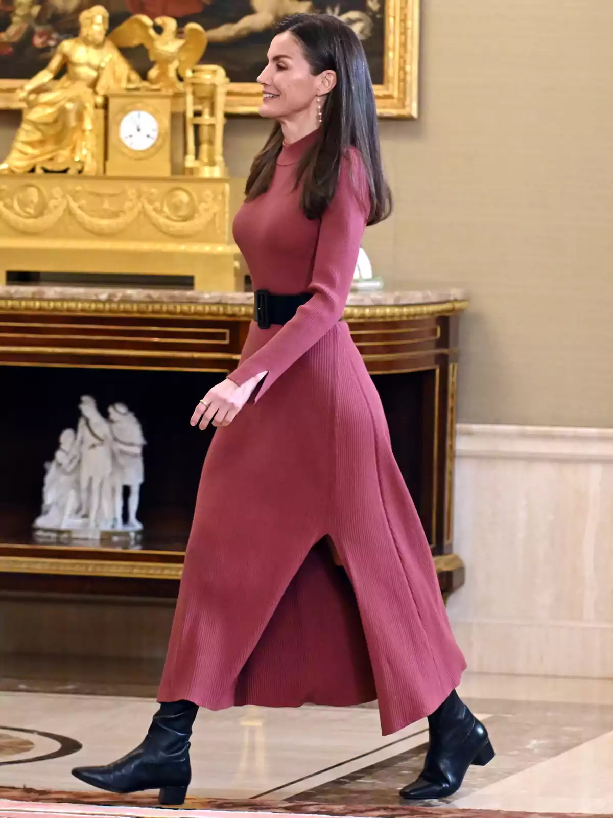 Queen Letizia walking in an elegant room, wearing a long dark pink dress and black boots, with a classic background decoration.