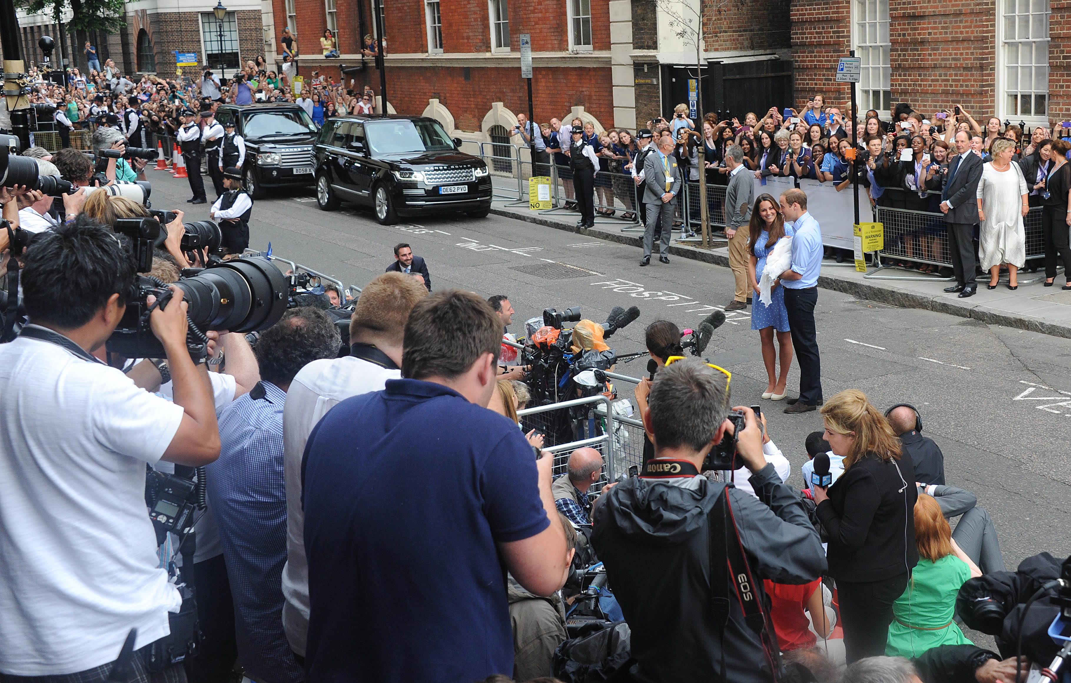 Kate Middleton and Prince William standing in front of a crowd of photographers with baby Prince George