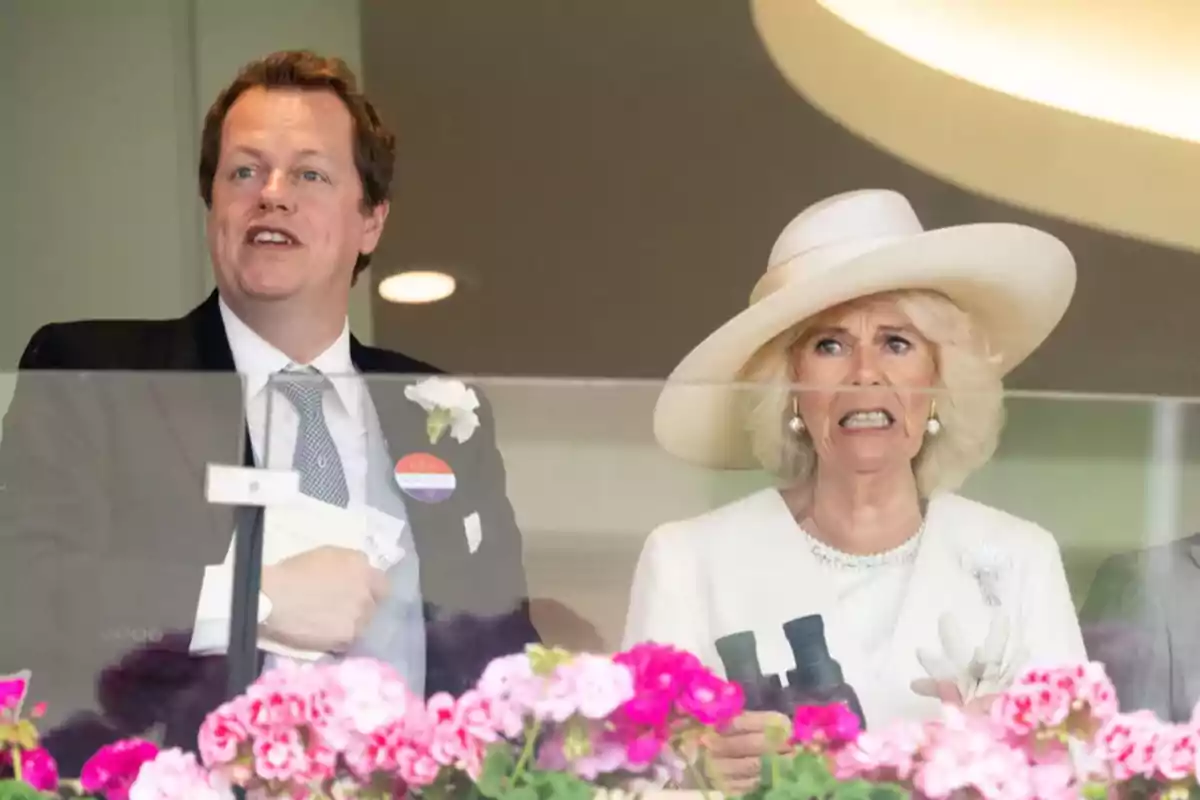 Elegantly dressed Tom Parker and Camila Parker watch something from a balcony decorated with pink flowers.