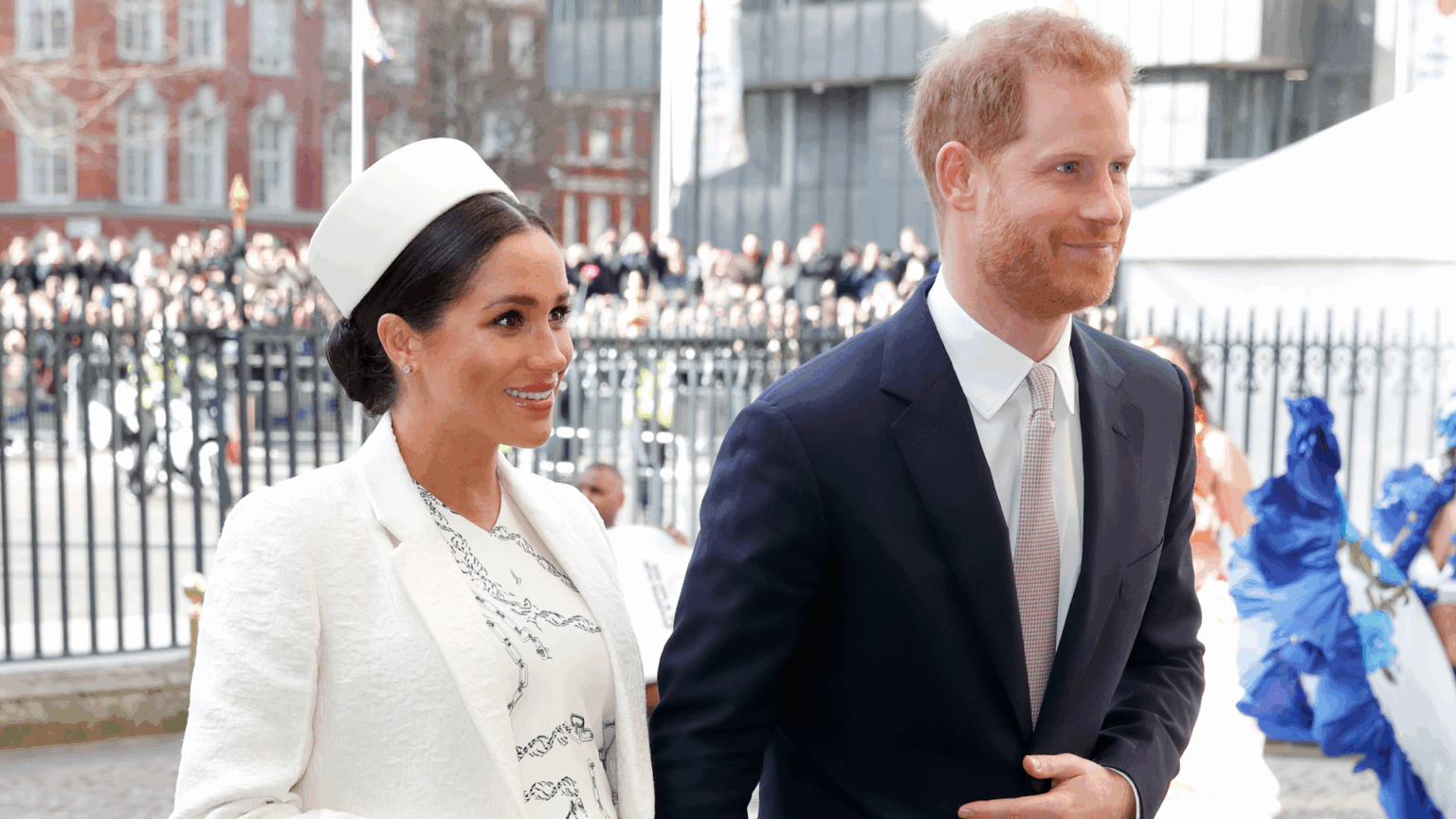 Meghan Markle Says One Aspect of Royal Life Left Her “Clinging Very Closely” to Prince Harry During Prince Archie Pregnancy Meghan Markle wearing a white hat and coat holding hands with Prince Harry at Westminster abbey