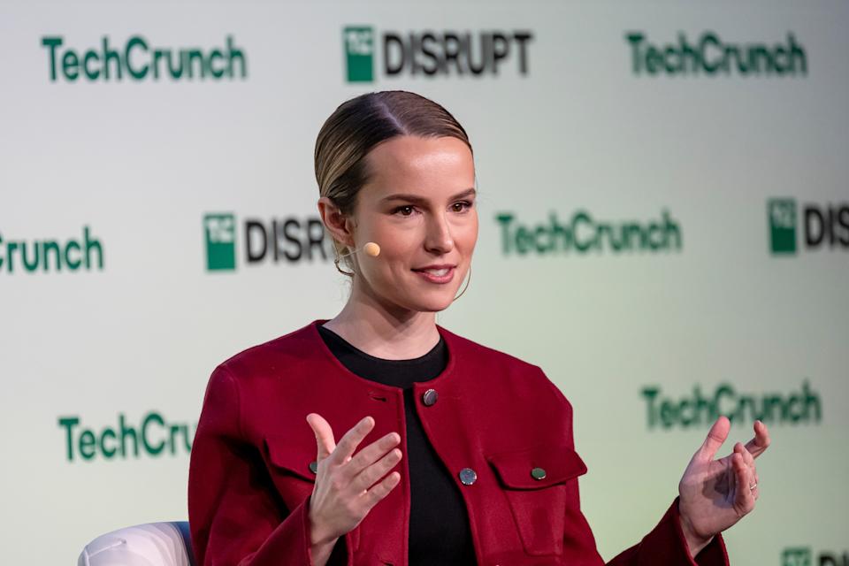 A person speaks onstage at TechCrunch Disrupt, gesturing with hands. They wear a sleek red jacket and have a microphone headset