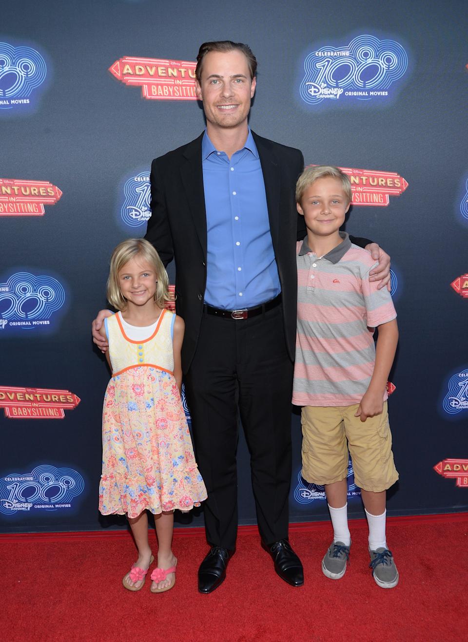 A man in a suit and open-collared shirt stands with two children on a red carpet at a Disney event