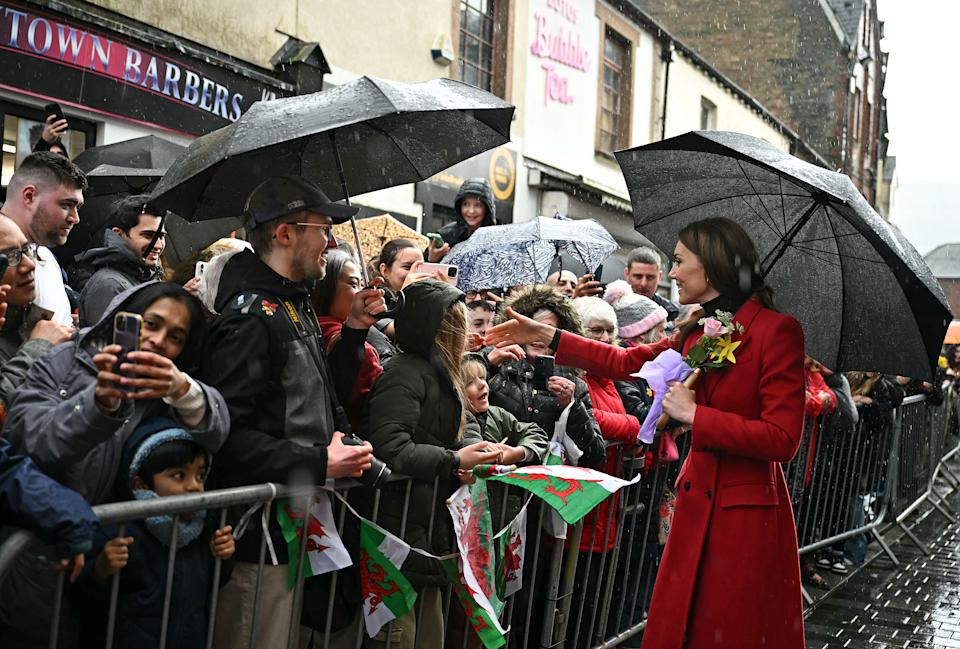 Kate Middleton wearing a red coat and holding a black umbrella talking to a crowd of fans