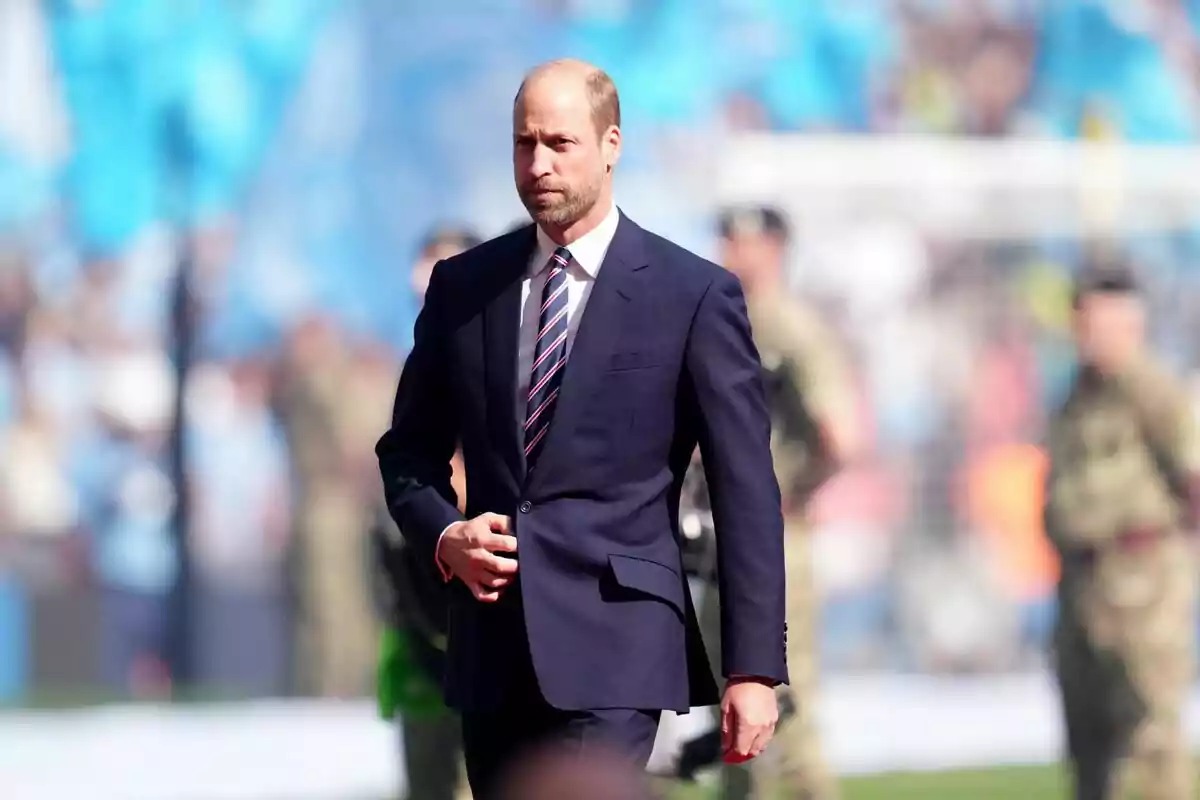 A man in a dark suit and tie walks at an outdoor event with uniformed people in the background.