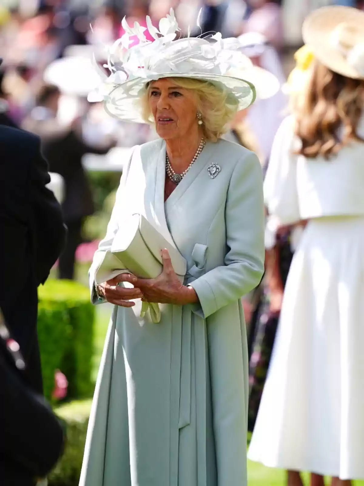 Queen Camilla elegantly dressed in a light blue coat and a hat decorated with white flowers, holding a handbag at an outdoor event Queen Camilla elegantly dressed in a light blue coat and a hat decorated with white flowers, holding a handbag at an outdoor event