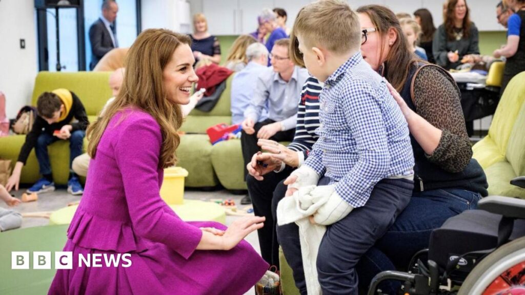 Princess of Wales wears a magenta skirt suit and is kneeling while chatting to a child and his carer. Other adults and children are in the room.