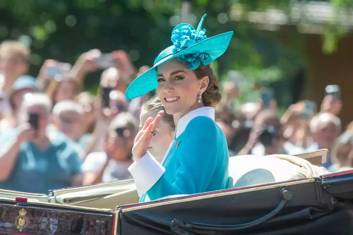 Woman in a blue hat smiles and waves from a carriage during an outdoor event with a crowd in the background