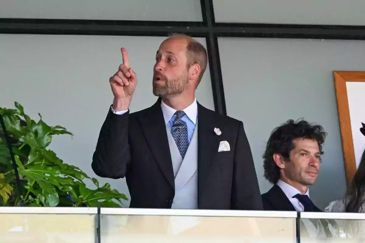 Man with a beard and dark suit raising his index finger while standing next to another person on an indoor balcony Man with a beard and dark suit raising his index finger while standing next to another person on an indoor balcony