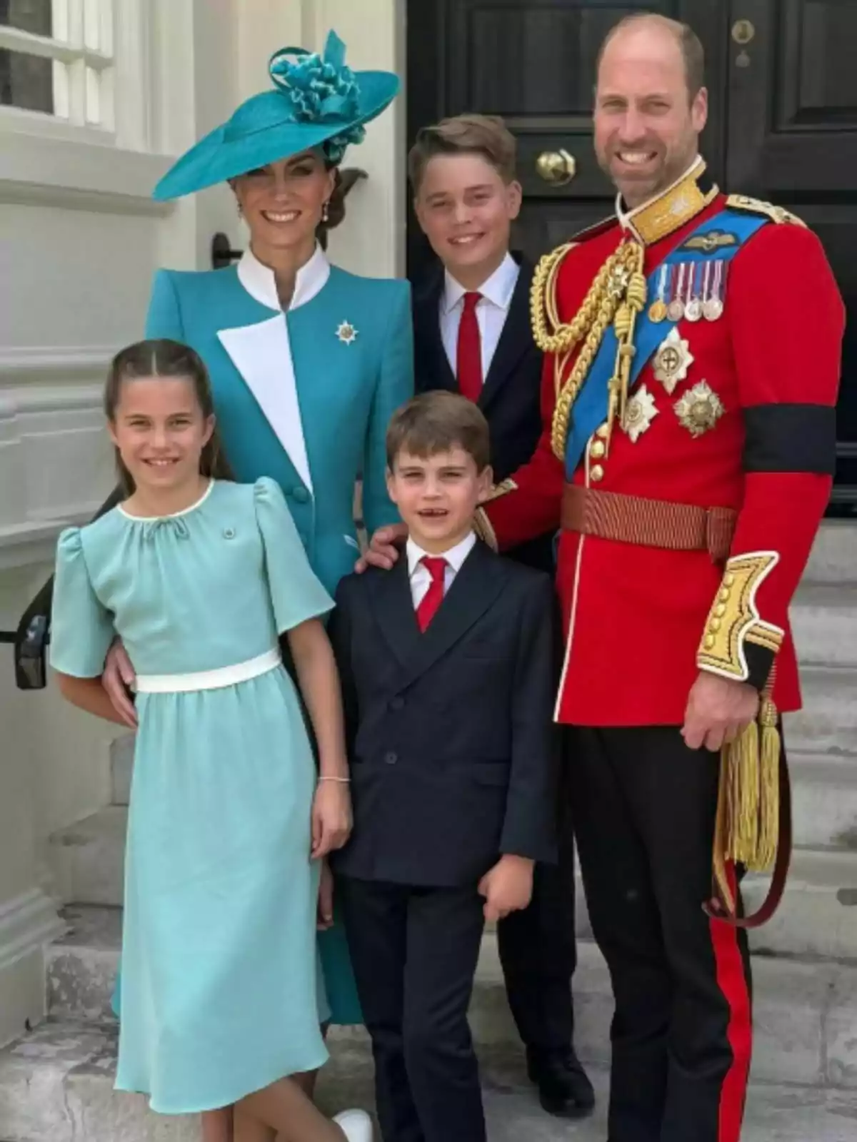 An elegantly dressed family poses smiling at the entrance of a building, with the man wearing a red uniform with medals and the woman a blue dress with a matching hat, accompanied by three well-groomed children.