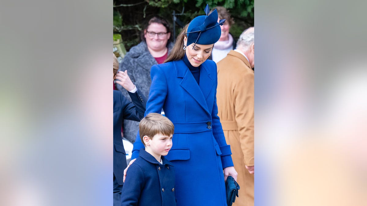 Kate Middleton in a blue coat dress touching Prince Louis' back