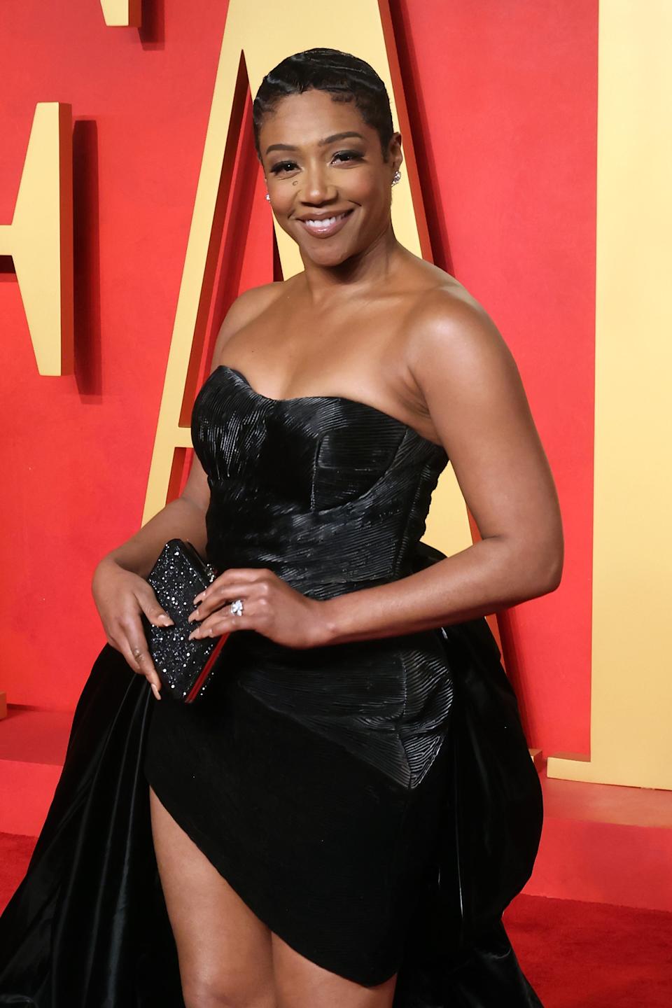 Tiffany Haddish in a strapless black gown on a red carpet, smiling and holding a clutch