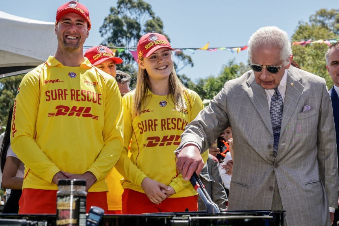 Two people look on as another person turns sausages on a barbecue.