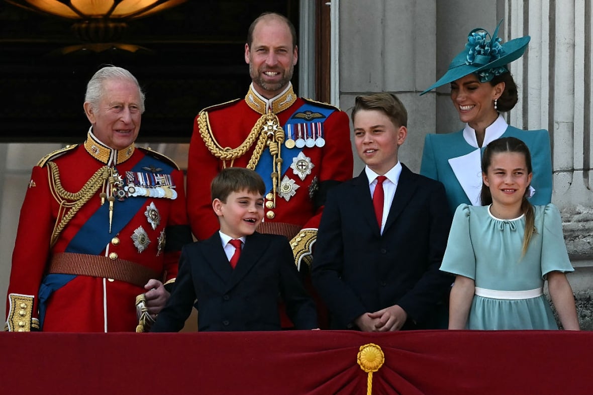 Six people stand on a balcony draped with a maroon-coloured covering in front of large open doors.