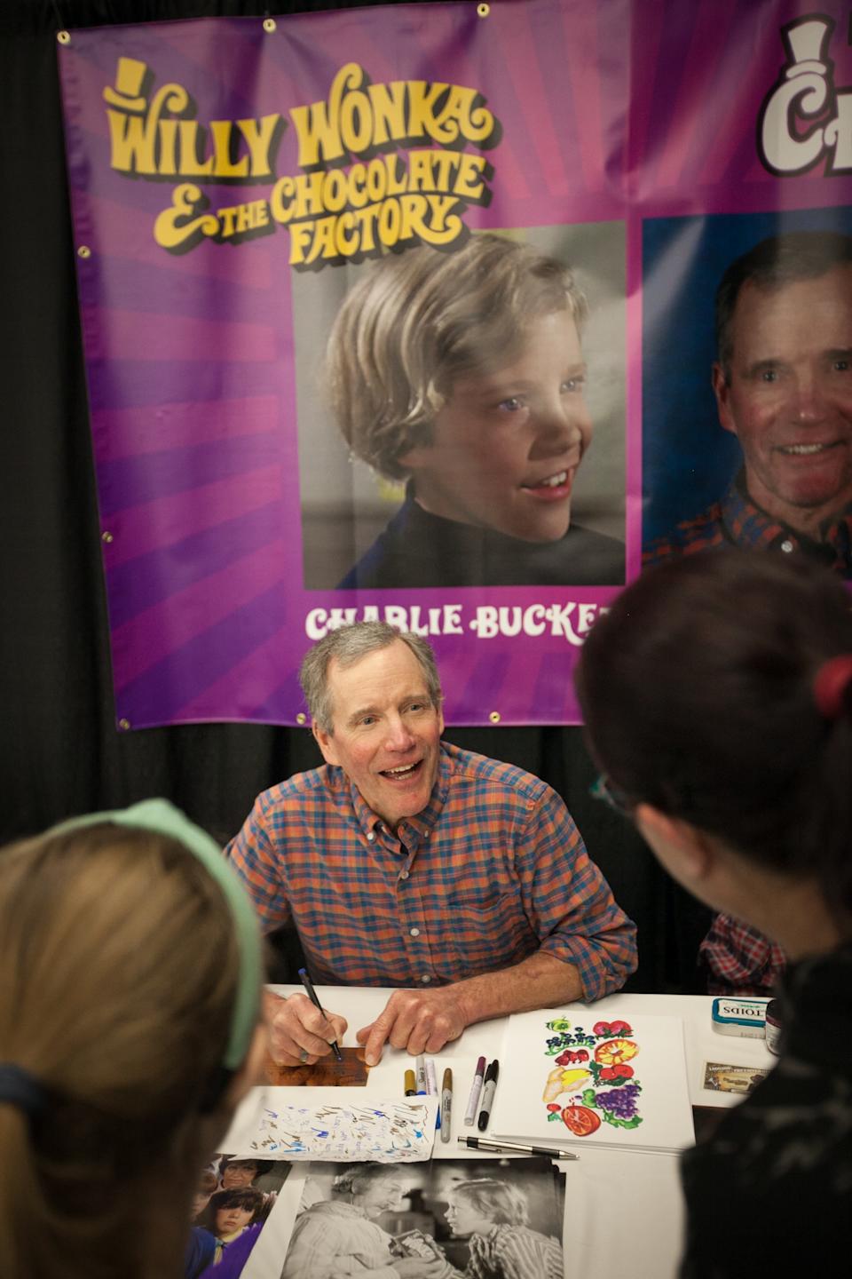 Person signing autographs at an event, sitting at a table with "Willy Wonka & the Chocolate Factory" posters