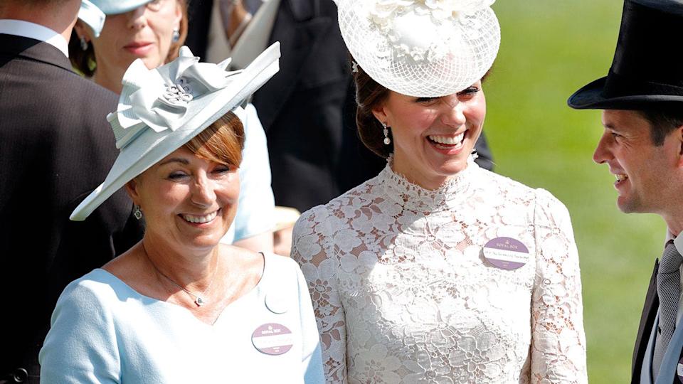 Kate Middleton smiling in a white lace floral dress and matching hat next to her mother.