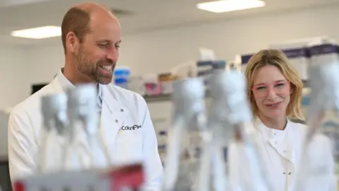 PA Media Prince William and Cate Blanchett are both smiling and standing inside a laboratory. They are both dressed in white lab coats with Prince William on the left and Cate Blanchett on the right, who is also 