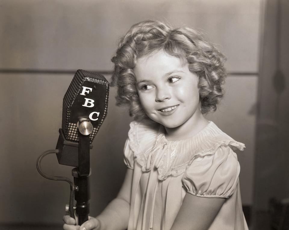 Shirley Temple with curly hair, wearing a dress, smiles while speaking into an old-fashioned microphone labeled "FBC."