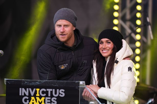 Prince Harry and Meghan Markle, both wearing dark woolly hats and large puffer jackets, hold each other's hands as they deliver a speech by a lectern. They are speaking at the 2025 Invictus Games.
