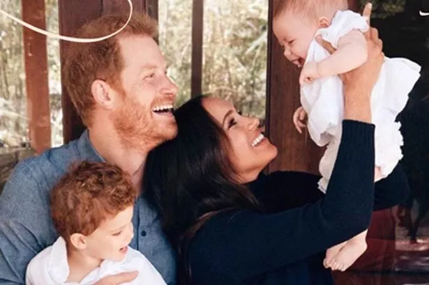 Prince Harry smiling with Prince Archie on his lap while Meghan Markle holds up Princess Lilibet 