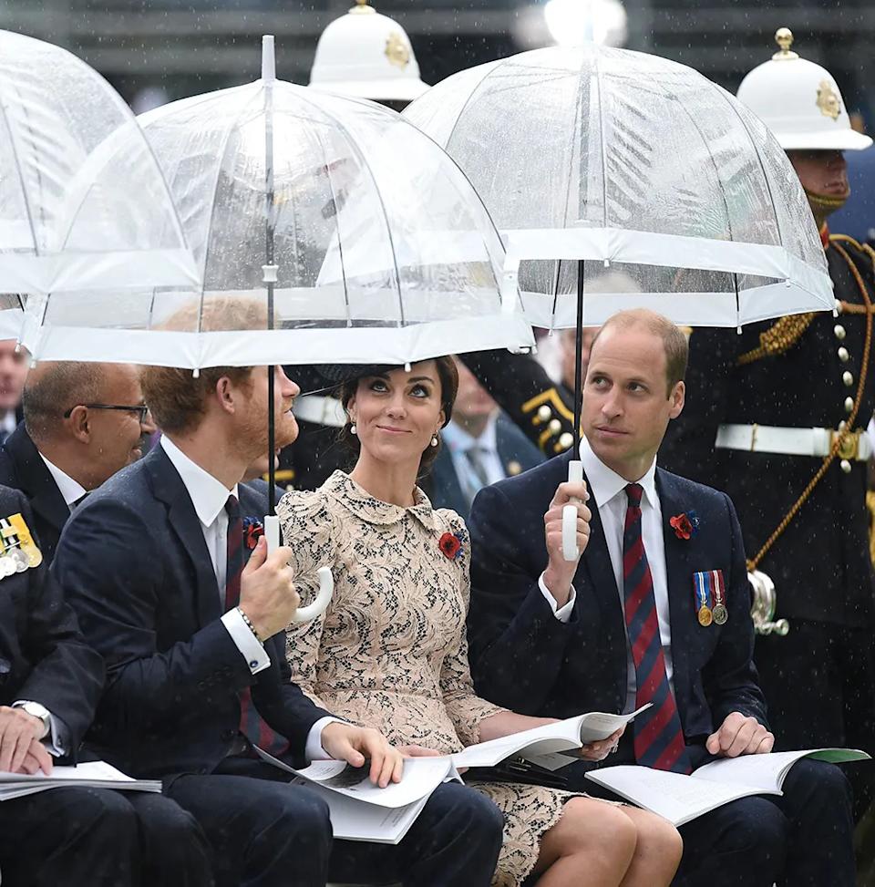 Prince Harry, Kate Middleton and Prince William sitting together in the rain with umbrellas