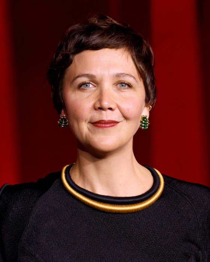Maggie Gyllenhaal with short hair wearing a black outfit with a gold collar and green earrings on a red carpet