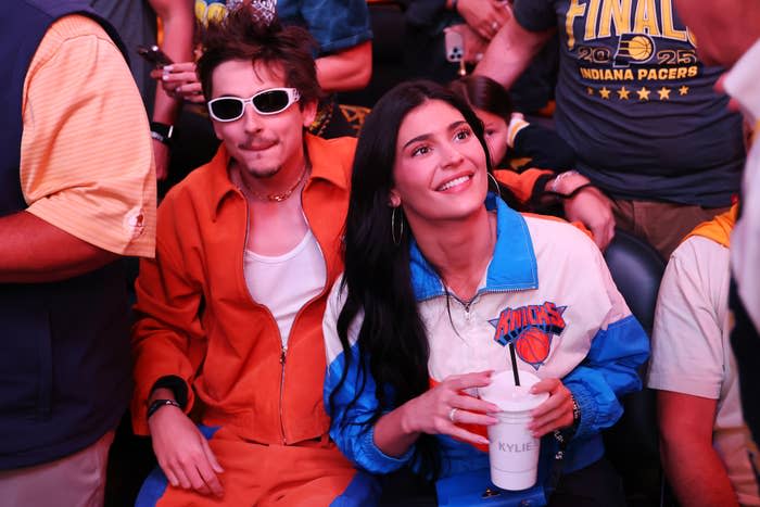 Timothee Chalamet and Kylie Jenner sit in a crowd, smiling. One wears a tracksuit. The other wears a basketball-themed jacket, holding a cup with "KYLIE" on it