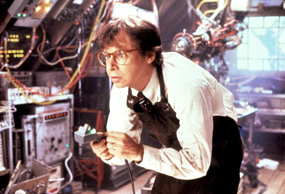 A person in a cluttered, high-tech lab, holding a remote. They wear glasses and a shirt with a tie, looking intently at the equipment