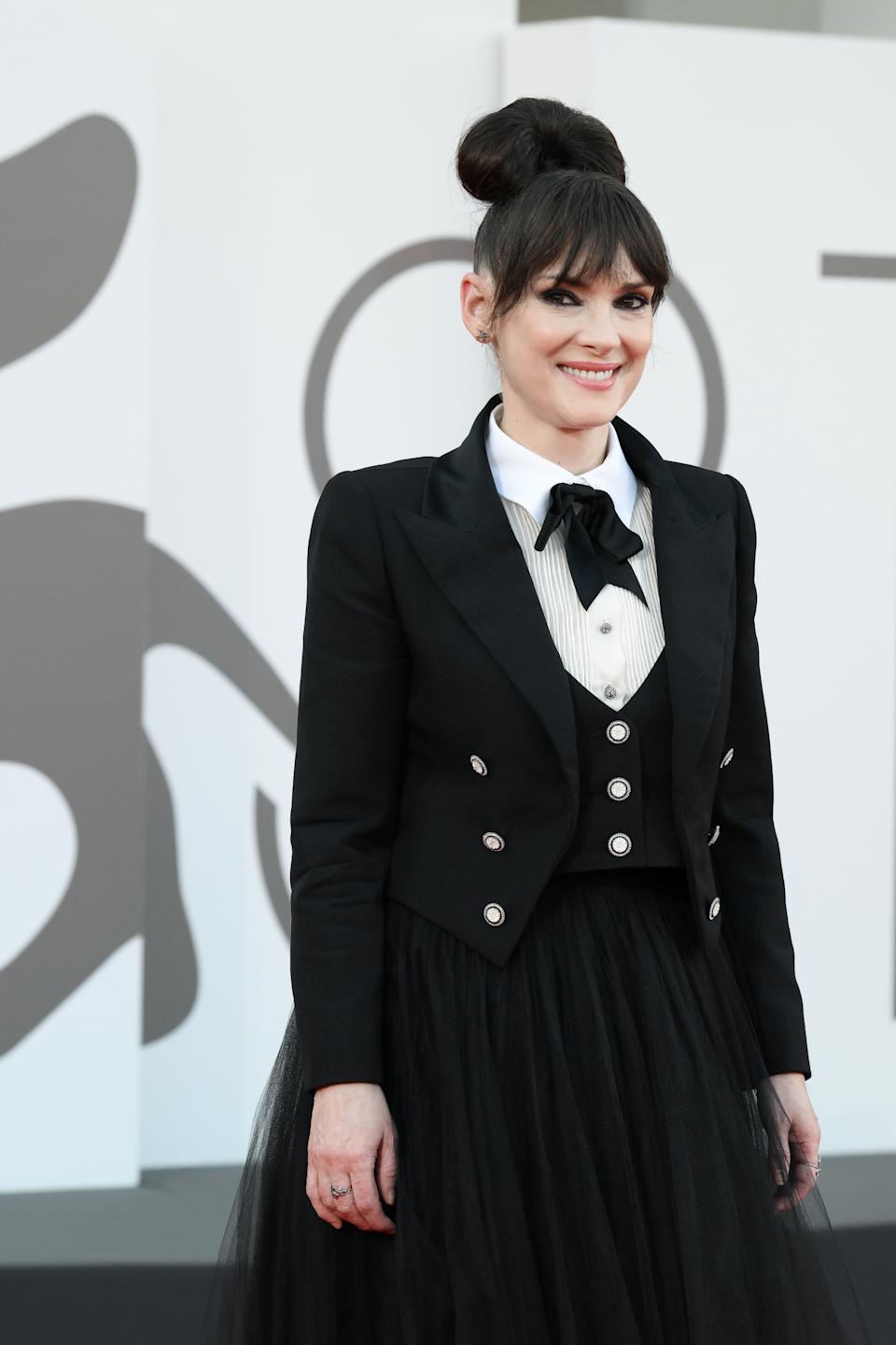 Winona Ryder on a red carpet wearing a chic tuxedo-style outfit with a tulle skirt and ribbon tie, smiling