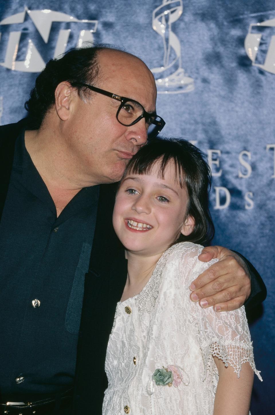 Danny DeVito in a dark suit lovingly embraces a smiling young Mara Wilson in a patterned dress, posing for photos at an event