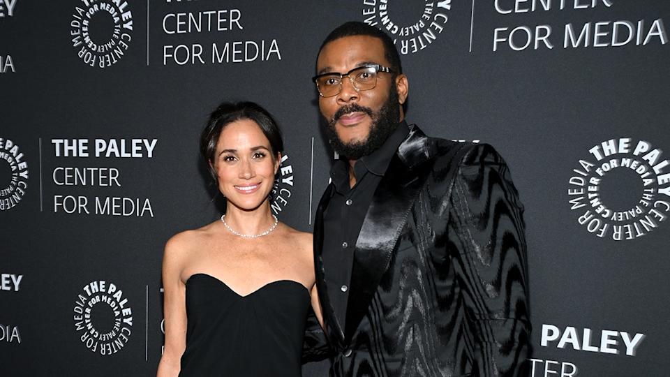 Meghan Markle wearing a strapless sweetheart cut black dress standing next to Tyler Perry wearing all black.