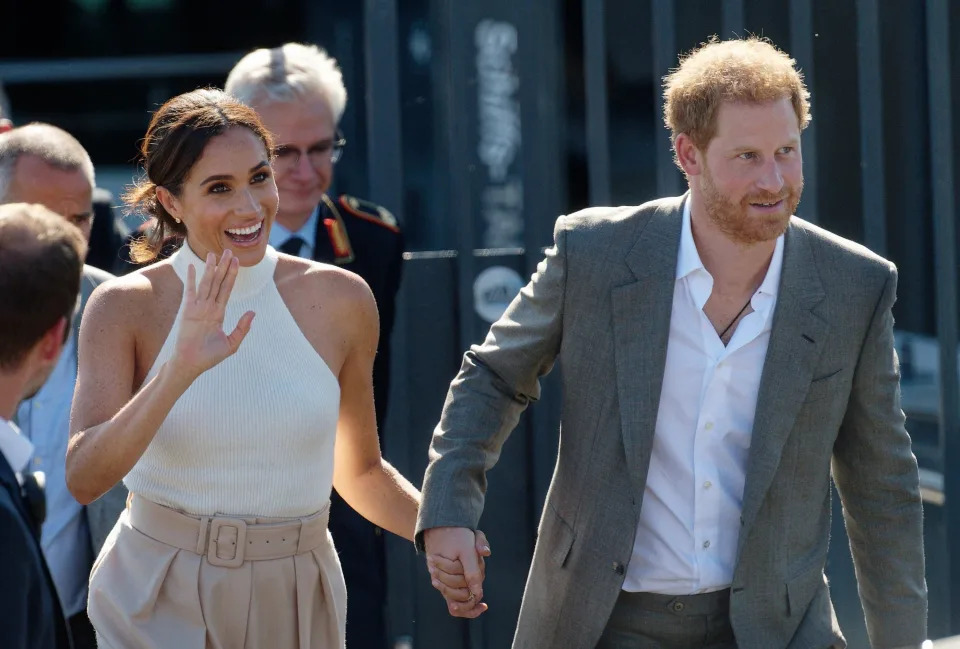 Meghan Markle waves in a white halter top and light pink pants while holding Prince Harry's hand in a grey suit